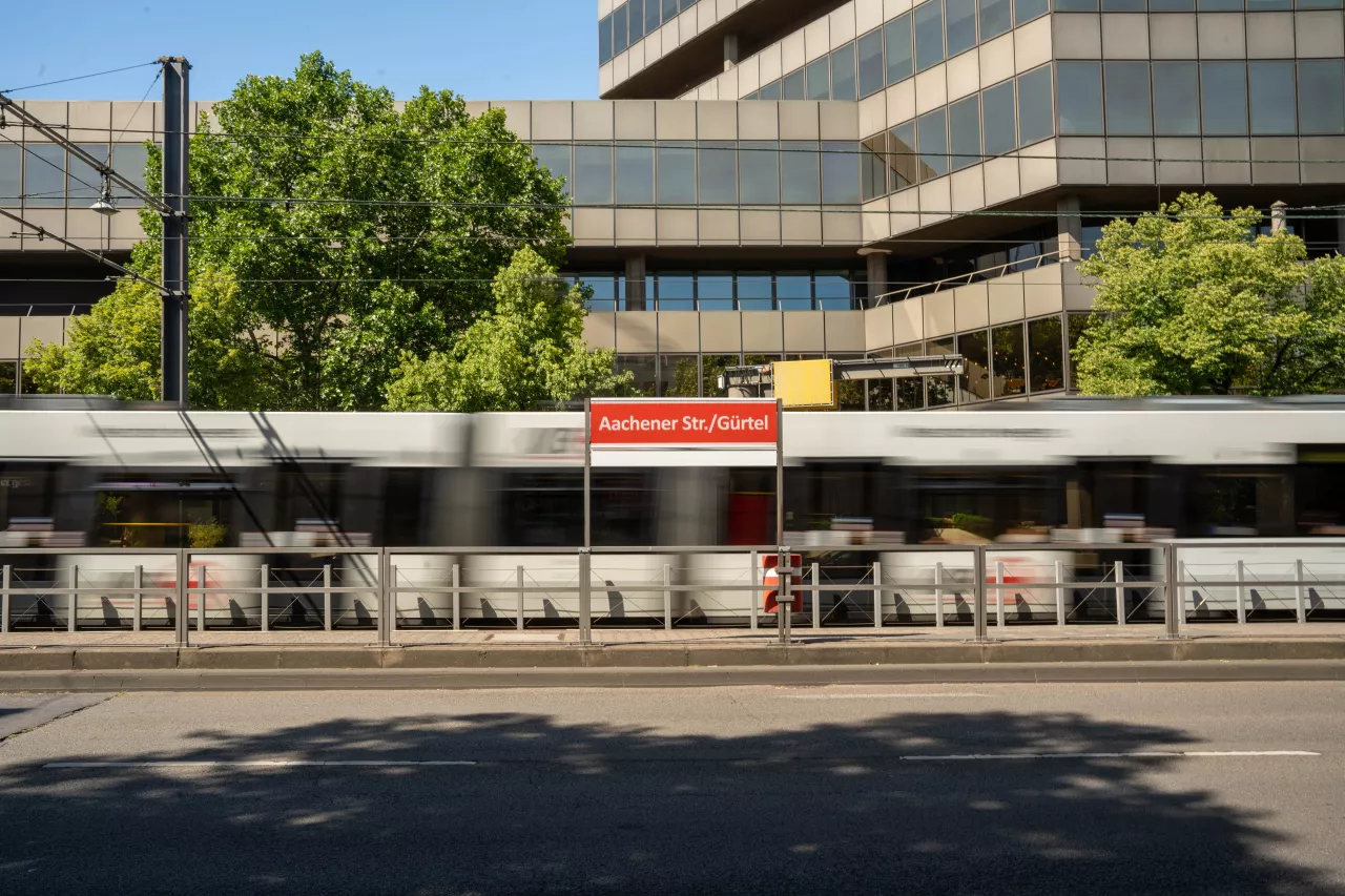 Haltestelle Aachener Straße/Gürtel Blick von der gegenüberliegenden Straßenseite auf das Haltestellenschild Aachener Straße/Gürtel mit fahrender Straßenbahn im Hintergrund