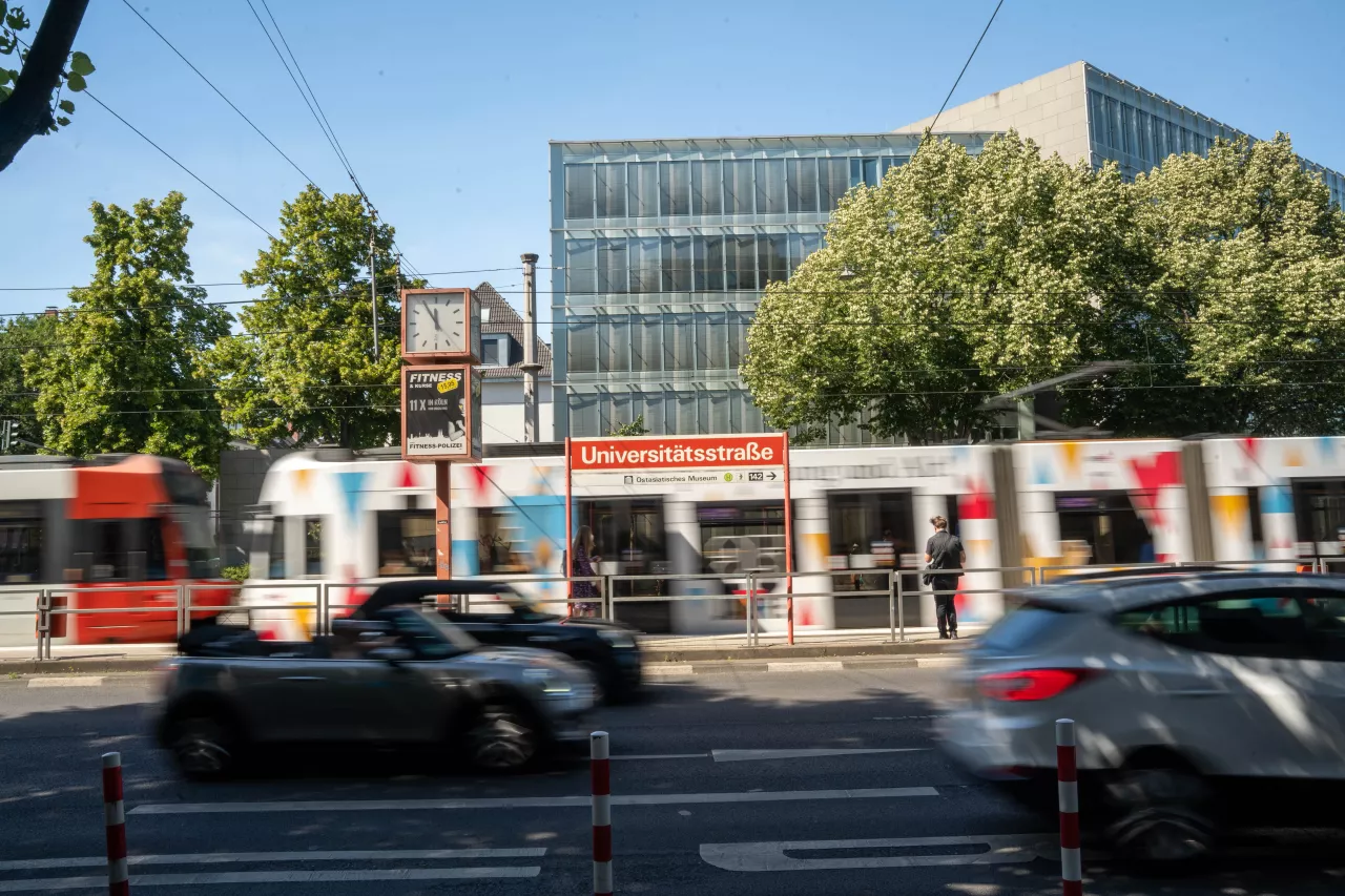 Haltestelle Universitätsstraße Blick von gegenüberliegender Straßenseite auf das Haltestellenschild Universitätsstraße mit Verkehr im Vordergrund und einer Straßenbahn im Hintergrund